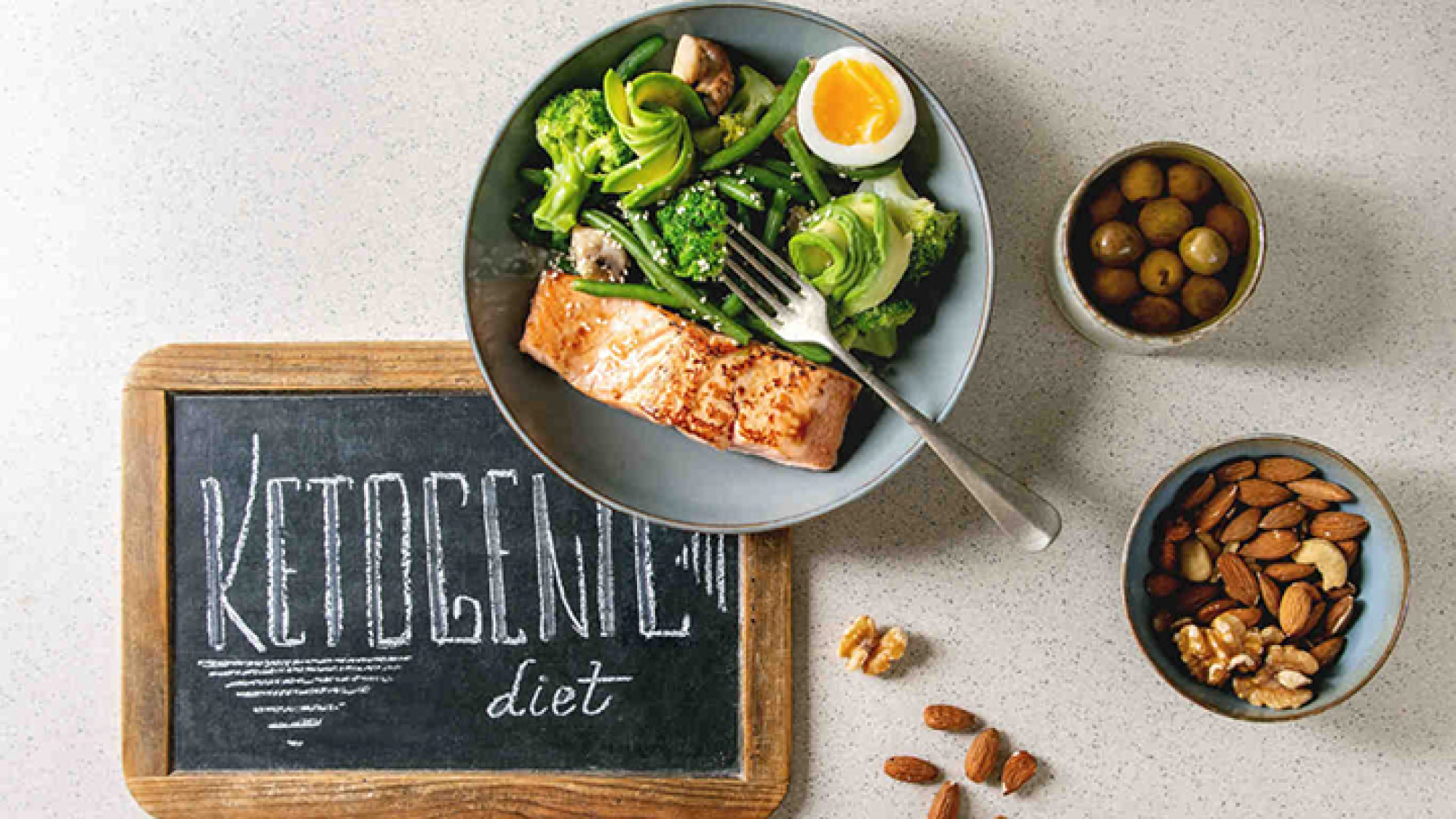 Ketogenic low carb diet dinner grilled salmon, avocado, broccoli, green bean and boiled egg in bowl served with olives, nuts and chalk board lettering over grey spotted background. Flat lay, space